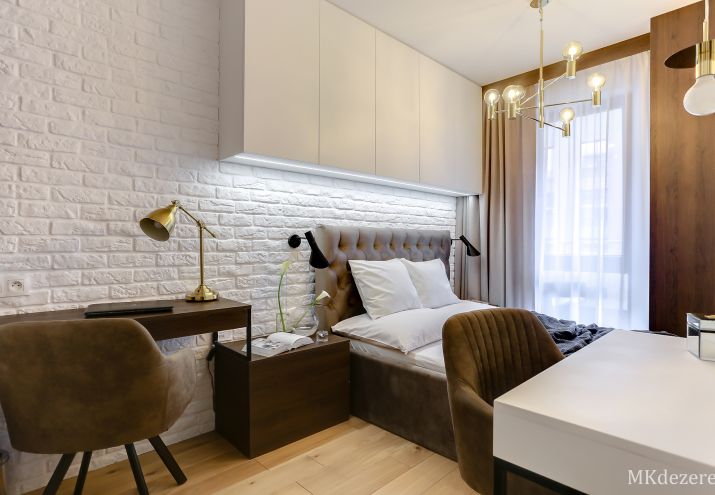 Aranżacja niewielkiej sypialni z białą cegłą i dodatkowymi szafkami nad łóżkiem.