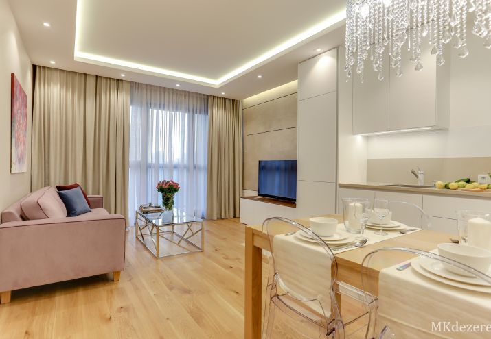 Aranżacja salonu z kuchnią i jadalnią. Salon w kolorze beżu, bieli oraz z ciepłym drewnem dębowym. W całym pokoju znajduje się dekoracyjny podwieszany sufit z wnęką na oświetlenie Led.
