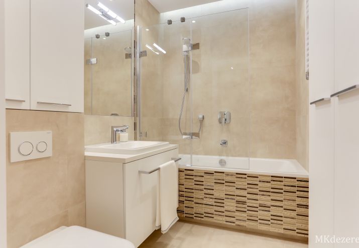 Aranżacja łazienki w beżach z mozaiką. W łazience znajduje się wanna z parawanem nawannowym składanym, duża umywalka oraz miska wc.