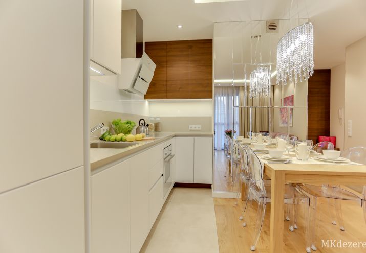 Aranżacja małego salonu z drewnianym stołem, duże lustro, blat w kuchni z kamienia, białe meble, lampa z kryształkami nad stołem.