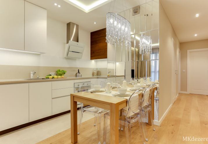 Stół w kuchni przy ścianie z przezroczystymi krzesłami. Na ścianie umieszczone dekoracyjne lustra w których odbija się żyrandol z kryształkami.